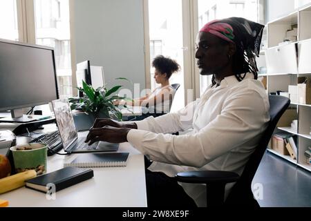 Male computer programmer using laptop while sitting on chair with colleague at office Stock Photo