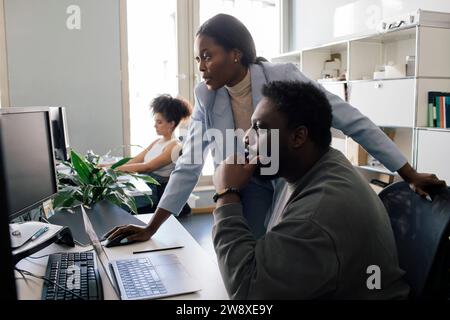 Male colleague with hand on chin sitting near female colleague assisting at office Stock Photo