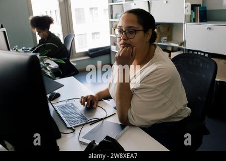 Businesswoman with hand on chin using computer while sitting at desk in office Stock Photo