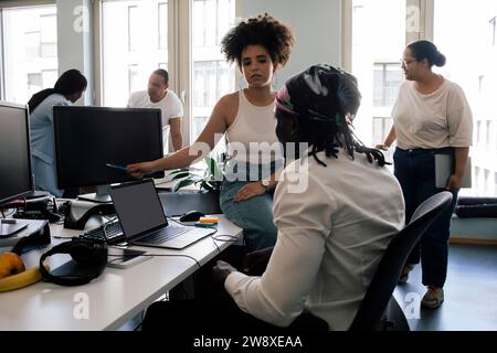 Female programmer explaining coding to colleague over computer while sitting at desk in office Stock Photo