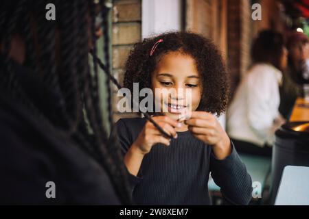 Smiling daughter with curly hair braided hair of mother while sitting at restaurant Stock Photo
