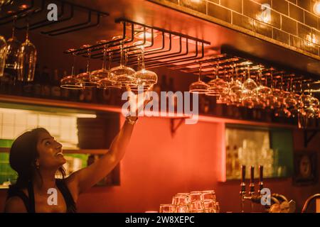 Smiling female bartender hanging cocktail glasses on rack while working at bar Stock Photo