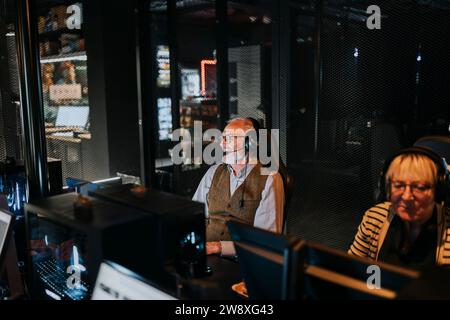 Elderly man wearing headset sitting by female friend playing video game on computer at gaming lounge Stock Photo