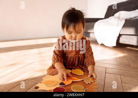 A cute little Asian girl is playing on the living room floor. A funny toddler in a brown dress attaches leaves to a hedgehog. Exercises for the develo Stock Photo