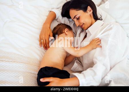 A cute baby sleeps with his attractive mother in a bed. A little boy falls asleep in the arms of his mother. A young smiling woman puts her sweet son Stock Photo