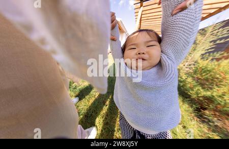 Close-up of a parent hands and a cute little Asian girl. Sweet Korean baby outdoors on a lawn with green grass. Mom and toddler are walking in the bac Stock Photo