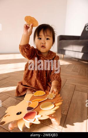 A cute little Asian girl is playing on the living room floor. A funny toddler in a brown dress attaches leaves to a hedgehog. Exercises for the develo Stock Photo