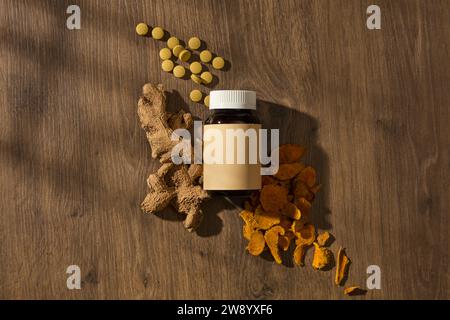 On the vintage wooden background, an unbranded bottle displayed with compressed capsules, dry ginger roots and dry slices or turmeric. Advertising sce Stock Photo