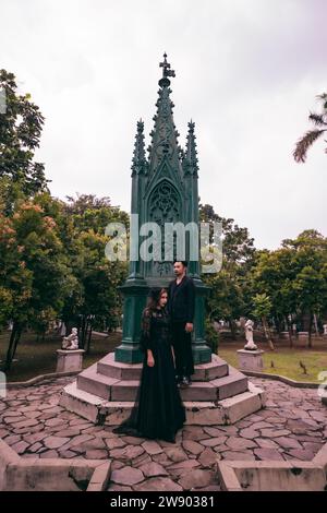 a couple of Asian teens standing near a spooky cemetery full of green trees in the afternoon Stock Photo