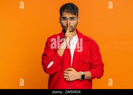 Shh be quiet please. Indian man presses index finger to lips makes silence gesture sign do not tells secret, stop talk gossip, confidential privacy. Arabian young guy isolated on orange background Stock Photo