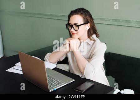 Connected and Smiling. Woman with Wireless Headphones Engages in Laptop Video Call, Watching and Talking. Happy Woman with Wireless Headphones Engages Stock Photo
