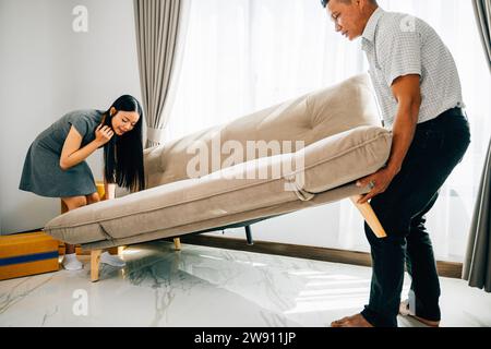 Asian couple smiles while moving a sofa in their new apartment Stock Photo