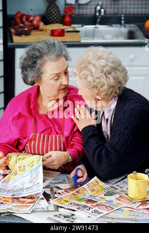 Two senior women best friends gossiping and clipping store coupons in a kitchen Stock Photo