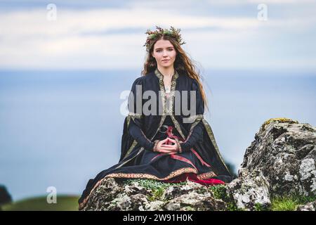 Young Female Pagan Witch in Winter Pine Floral Crown Mountain Peak Top Bay View Sitting Rocks Afternoon Solstice Day Stock Photo