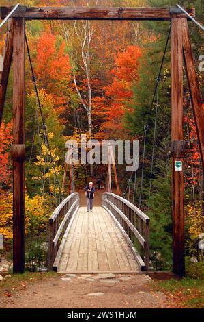 An active senior walks across a pedestrian suspension bridge to view the fall colors of New Hampshire's White Mountains on an autumn day n New England Stock Photo