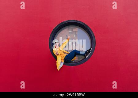 Woman in yellow rain coat sitting in porthole making victory sign Stock Photo