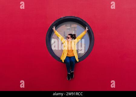 Woman in yellow rain coat sitting in porthole making victory sign Stock Photo