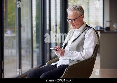 Senior man sitting on chair at home using mobile phone Stock Photo