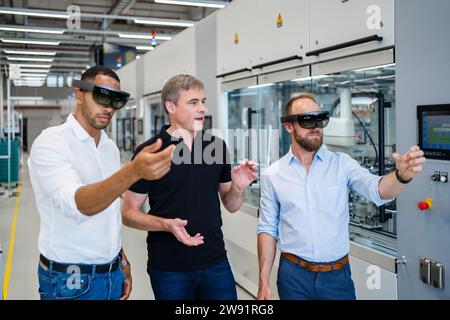 Two technicians wearing augmented reality glasses in a factory and interacting with colleague Stock Photo