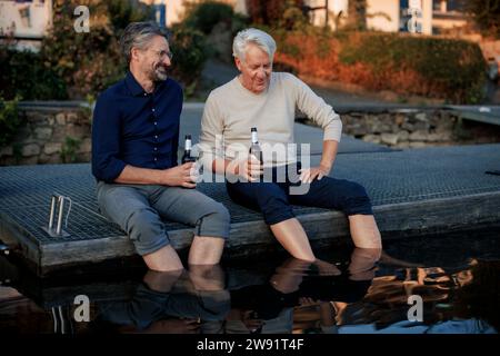 Happy senior friends sitting on pier with beer bottles Stock Photo