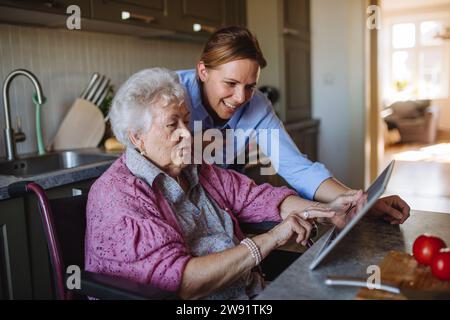 Happy home caregiver teaching tablet PC to senior woman at table Stock Photo