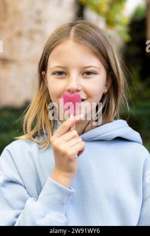 Smiling blond girl holding heart shaped lollipop candy over mouth Stock Photo