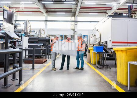Young engineers with protective workwear discussing over blueprint at industry Stock Photo