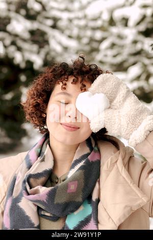 Smiling woman covering eye with heart shaped snow in winter Stock Photo