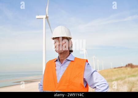 Contemplative engineer wearing hardhat standing in front of wind turbines Stock Photo