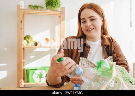 Smiling redhead woman putting plastic bottles in mesh bag at home Stock Photo