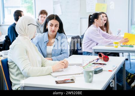 Woman discussing with friend in hijab sitting at desk in classroom Stock Photo