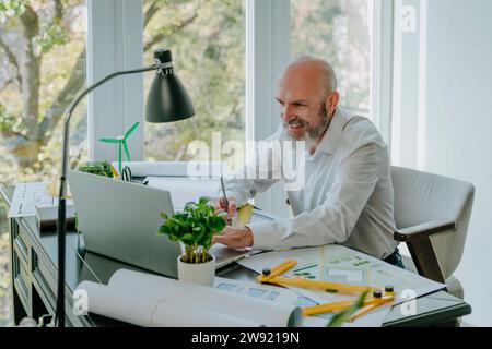 Happy mature architect working on laptop with blueprints at desk Stock Photo