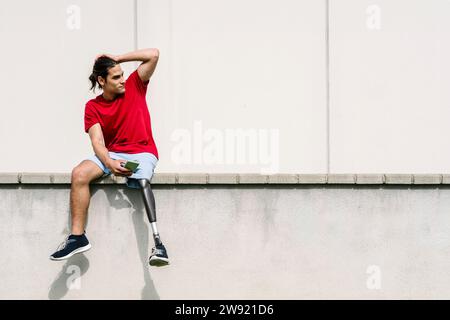 Young man with disability holding smart phone and sitting in front of wall Stock Photo