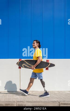 Smiling young man holding skateboard and walking with prosthetic leg in front of wall Stock Photo