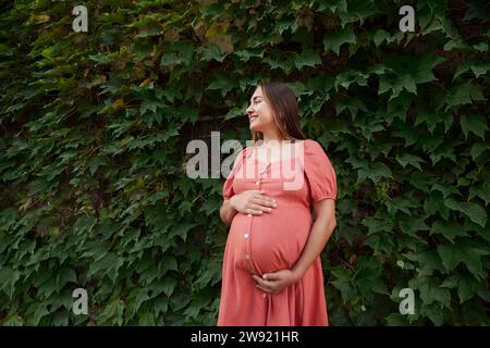Happy pregnant woman wearing dress and standing in front of plants Stock Photo