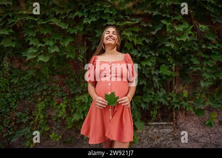 Happy young pregnant woman holding maple leaf in front of plants Stock Photo