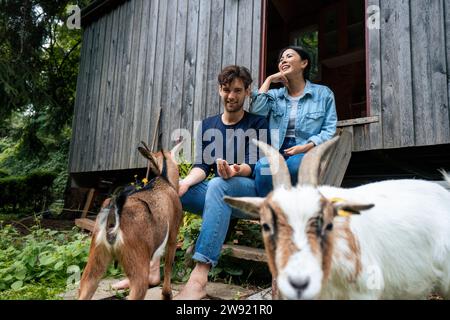 Happy couple sitting on steps with goats near log cabin Stock Photo