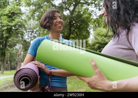 Happy friends holding exercise mats and talking to each other at park Stock Photo