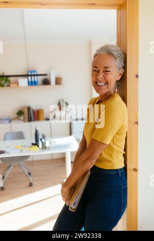Happy mature businesswoman holding file folder and leaning on doorway in office Stock Photo