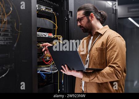 Engineer holding laptop and examining cables in server room Stock Photo