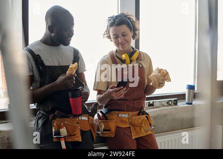 Smiling construction worker using smart phone and having lunch with coworker at site Stock Photo