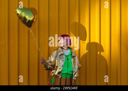 Smiling senior woman holding heart shaped balloon in front of yellow wall Stock Photo