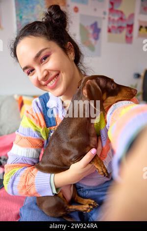 Smiling young woman holding dog and taking selfie at home Stock Photo