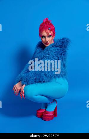 Non binary person with pink hair crouching against blue background Stock Photo
