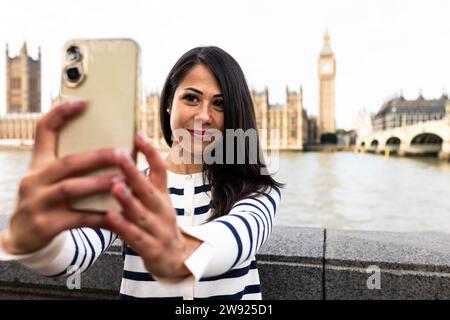 Smiling woman taking selfie with Big Ben and Houses of Parliament through smart phone in London city Stock Photo