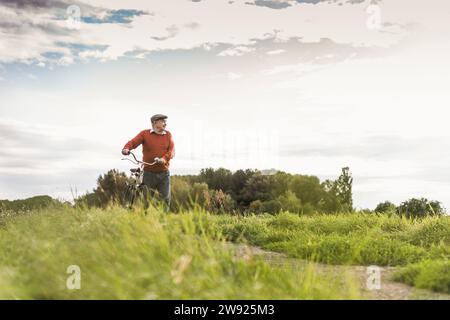 Senior man walking with bicycle on footpath at field under cloudy sky Stock Photo