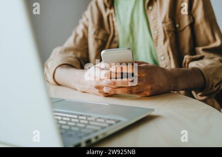 Man text messaging through smart phone by laptop on table at home Stock Photo