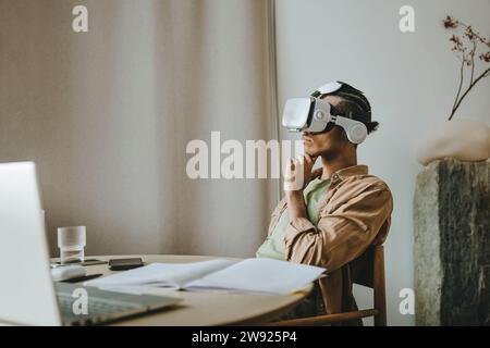 Young man wearing VR headset sitting with hand on chin at table Stock Photo