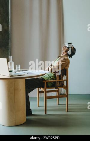 Young man wearing VR simulator sitting with arms crossed and head back at table Stock Photo