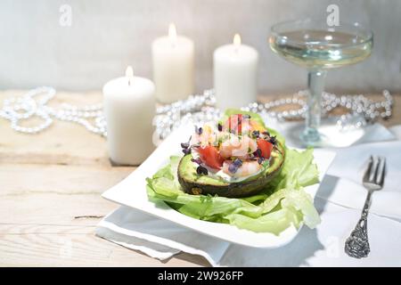 Salad from shrimps, tomatoes and mayonnaise filled in a halved avocado on a light rustic wooden table, wine glass, white candles and festive decoratio Stock Photo